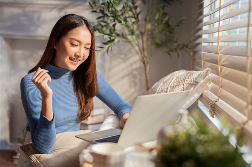 Smiling freelancer Asian female adult woman sitting on floor working with laptop on wooden bench next to living room window morning casual lifestyle ,Working From Home in Cozy Sunny Atmosphere