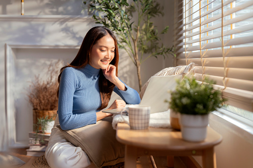 Smiling freelancer Asian female adult woman sitting on floor working with laptop on wooden bench next to living room window morning casual lifestyle ,Working From Home in Cozy Sunny Atmosphere