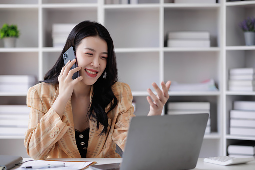 A woman is talking on her cell phone while sitting at a desk with a laptop. She is smiling and she is in a good mood