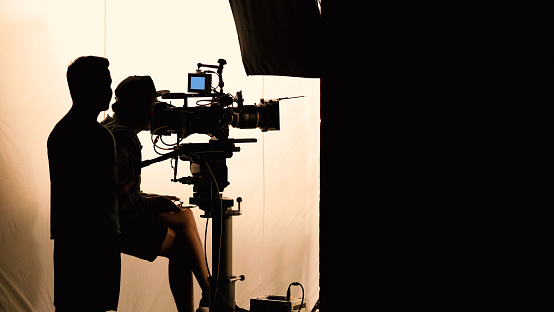 Video production behind the scenes which film crew team in silhouette shooting or recording tv movie commercial with professional equipment such as high definition 4k camera with monitor and tripod or gimbal rig in big studio set.