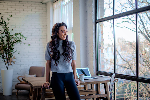 A young african american woman standing at the window and looking thoughtfully. Long haired female wearing casual clothes.