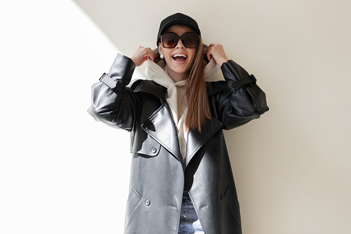 Happiness young beautiful fashion woman hipster with happy emotions with trendy sunglasses and black hat in leather coat and hoodie wears a hood have fun enjoy near a wall on the street in sunlight