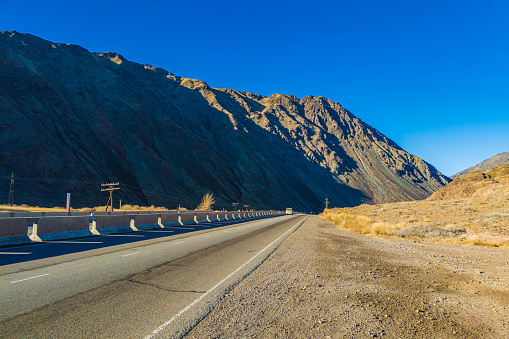 An empty highway stretches through a natural landscape with mountain in the background, showcasing the asphalt road surface as it winds through the hilly terrain.
