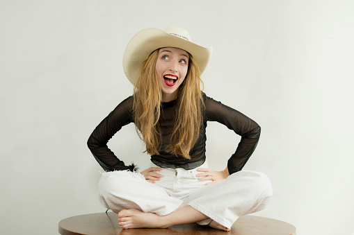 A pretty young blonde woman in white jeans and lacy black top poses in a studio setting wearing a straw cowboy hat.