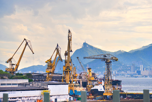 Crane lifting heavy cargo into a container at a busy port in Rio de Janeiro, with Christ the Redeemer in the background.