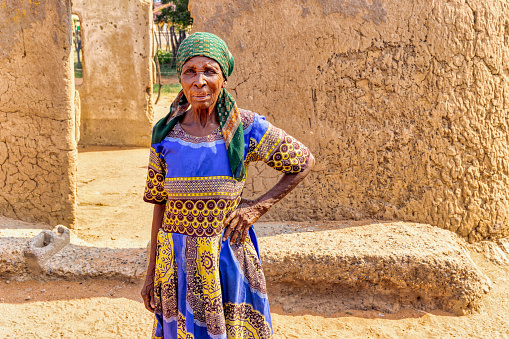 village old african woman standing in front of a ruined mud house
