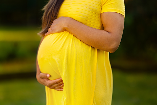 A caring pregnant woman in a form fitting yellow dress cradles her belly in a garden with trees and flowers.