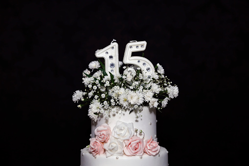 Three-tiered white wedding cake decorated with flowers, green leaves, figures of bride and groom on a white wooden background.