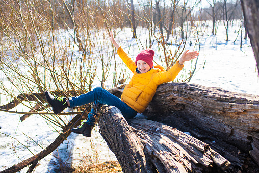 Happy 11 year old boy in yellow jacket and red hat sitting on fallen tree in spring forest against a background of snow