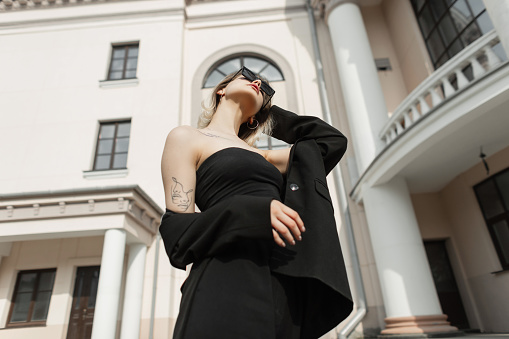 Cool glamorous young woman model with sunglasses in fashion black business elegant clothes with blazer, top and pants walking in the city near a vintage building