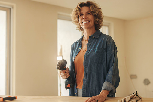 Mature, happy woman holds a power drill while working on a home renovation project in her kitchen. She stands with a smile, ready to upgrade and remodel her house with DIY expertise.