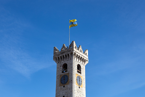 View of Clock Tower (Torre Civica or Torre di Piazza) and Trento flag in a blue sky sunny day; Trento, Trentino-Alto Adige, Italy
