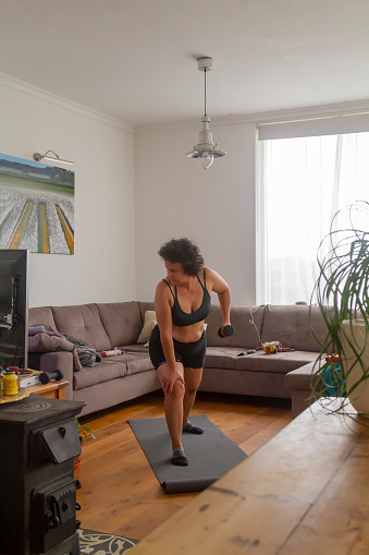 Mid Adult Mature senior Woman doing pilates, yoga and strength training exercises workout at home on a mat in her living room following the directions from a personal trainer by tv.

Her cat keeping her company and trying to play around her, wearing a black top shirt and black stretch exercises pants at morning time.