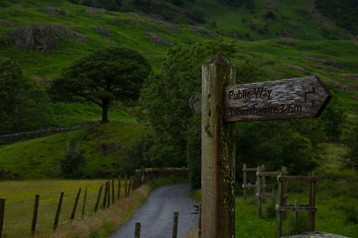 An old wooden signpost points the way to Thirlthwaite on a serene country lane in the Lake District, flanked by lush fields and dry stone walls. Countryside crossroad marked with a directional sign.