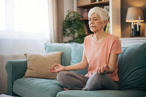 Senior woman with gray hair sitting on the sofa with legs crossed and meditates.