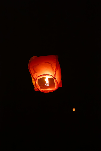 A single glowing orange sky lantern rises against a dark night background, surrounded by faint lights of other lantern