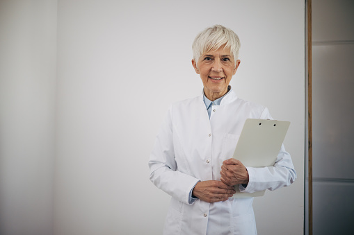 Senior female medical doctor standing in medical cabinet wearing uniform and holding fascicle.