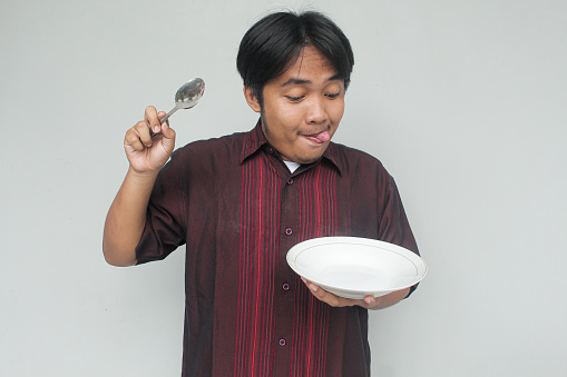 Asian young man looking to empty eating plate and holding spoon with funny expression.