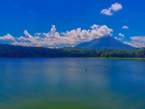 An aerial view of a clear blue lake with a dramatic, clouds capped mountain in the background. Evergreen trees line the shore near Ranu or Lake Klakah, Lumajang, East Java, Indonesia.