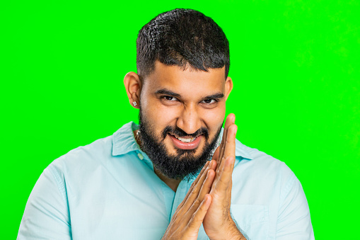 Sneaky cunning Indian young man with tricky face gesticulating and scheming evil plan, thinking over devious villain idea, cheats, jokes, pranks. Arabian guy isolated on green chroma key background