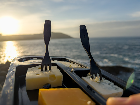 a picturesque scene of a cheese tasting session against the backdrop of a stunning sunset by the sea. The focus is on a black tray holding an assortment of cheeses, each pierced with a sleek black fork, standing out against the warm glow of the sun reflecting off the ocean. The image evokes a sense of relaxation and the simple pleasures of enjoying fine food with a view.