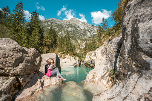 Two diverse female friends sit on rocks by a mountain stream, enjoying a sunny day outdoors on a hike.