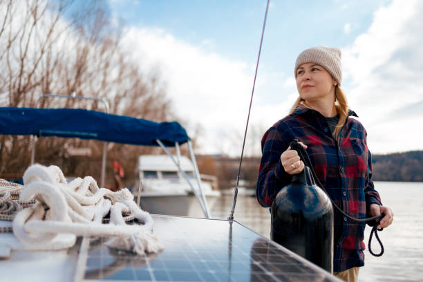A woman in a knitted hat and plaid shirt is carefully attaching a black fender to the side of a boat to protect it from bumping into the dock.