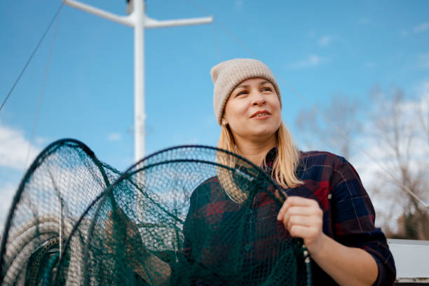 A woman in a warm beanie looks into the distance as she holds a green fisherman's net aboard a boat, with the clear blue sky above and the mast of the boat standing tall in the background.