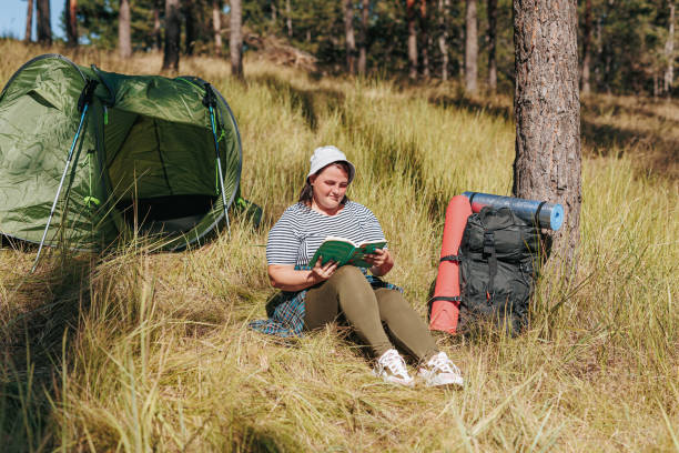 istock Journey of Pages Overweight Hiker Relaxing Read by Backpack and Tent 2110620049