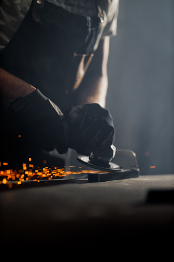 A skilled metalworker in safety glasses and gloves is intensely grinding a piece of metal, with sparks flying around in a dimly lit workshop.