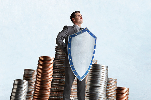 A man holding a medieval shield stands confidently and protectively in front of several stacks of coins.