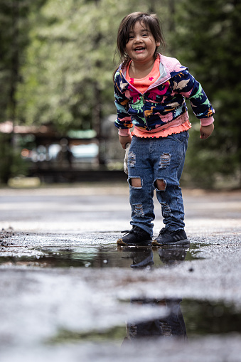 A young girl is standing in a puddle of water, wearing a blue and pink jacket. She is smiling and she is enjoying herself