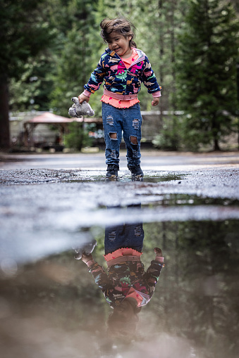 A young girl is standing in a puddle of water, holding a stuffed animal. The scene is playful and lighthearted, with the girl enjoying her time outdoors