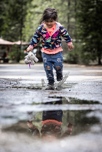 A young girl is playing in the rain, splashing water and holding a stuffed animal. The scene is playful and joyful, with the girl enjoying the moment and the rain