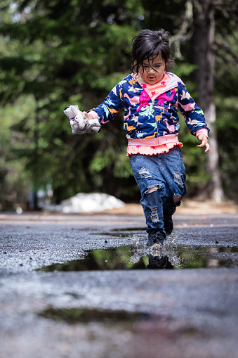 A young girl is playing in the rain, splashing in puddles and holding a stuffed animal