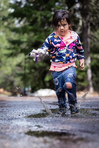 A little girl is running through a puddle of water, holding a stuffed animal. The scene is playful and lighthearted, with the girl enjoying her time outdoors