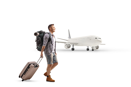 Male tourist with a backpack and a suitcase walking in front of a plane isolated on white background