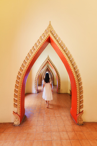 Rear view of asian woman walking in traditional golden arch door with Thai style carve decoration in corridor at Wat Tham Sua, Kanchanaburi, Thailand