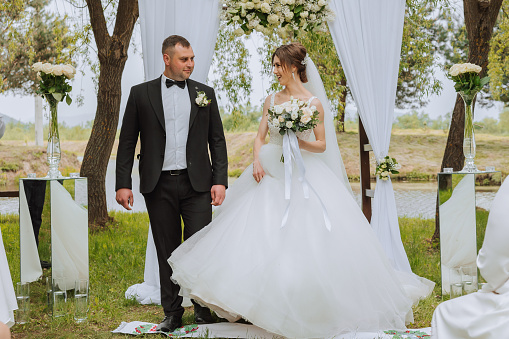 A groom in a black suit and a bride in a voluminous dress stand near a white arch decorated with flowers during a wedding ceremony. The bride is holding a bouquet. Spring wedding