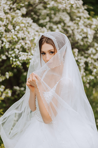 Red-haired bride in a lush dress with an open bust, posing wrapped in a veil, against the background of flowering trees. Spring wedding in nature.
