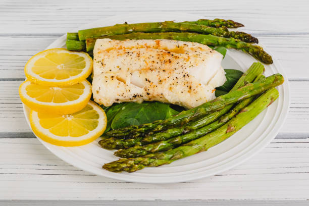 istock Cod fillet baked with garlic butter sauce served with fresh spinach and roasted asparagus on a white plate close-up 2110872265