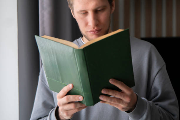 istock A focused young man wearing a casual grey sweater is deeply absorbed in reading a green hardcover book while seated comfortably by a window sill. 2110890281
