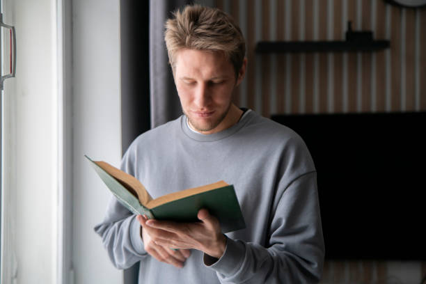 istock A focused young man wearing a casual grey sweater is deeply absorbed in reading a green hardcover book while seated comfortably by a window sill. 2110890283