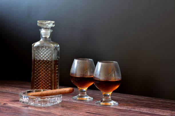 two glasses and a crystal decanter of cognac on a wooden table, next to a glass ashtray with a cigar. - decanter crystal carafe glass imagens e fotografias de stock