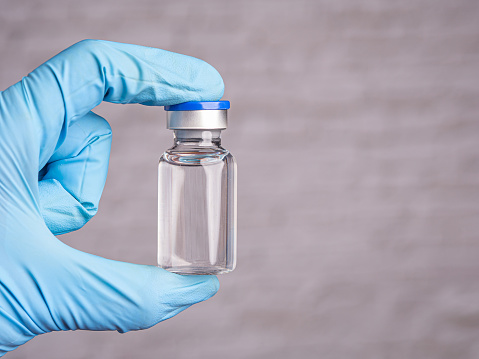 The doctor's hand in blue gloves holds a vaccine vial on a gray background. Space for text.