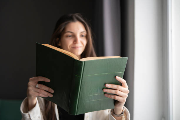 istock A woman stands by a large window, bathed in natural light, deeply focused on reading an open book. 2111016749