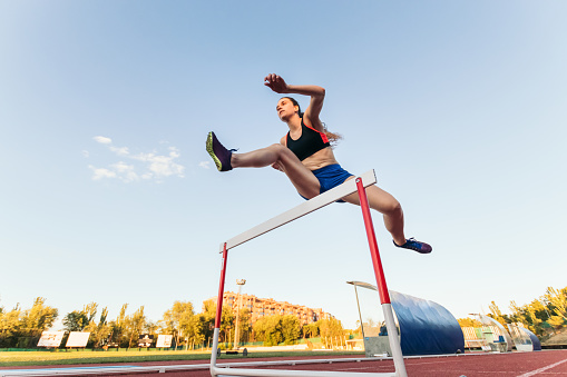 A strong woman in sports attire training for an obstacle race. She is jumping hurdles on an athletics track.