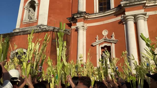 Catholics celebrate Palm Sunday before Easter by waving coconut palm leaves in commemoration of Jesus' triumphal entry into Jerusalem, with the church tower in the background.