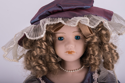 The face of a porcelain doll wearing a vintage lace hat and long curly hair. Close-up.