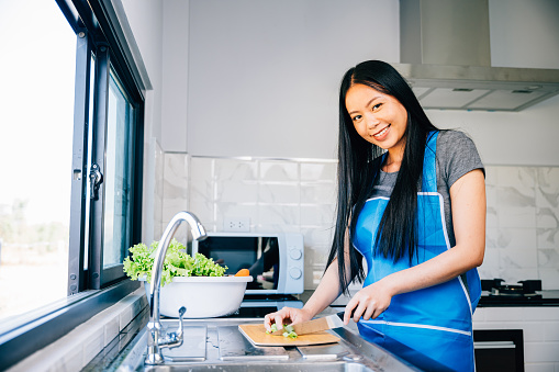 A smiling woman in an apron prepares a healthy dinner cutting vegetables for a delicious salad in her kitchen. Close-up of a cheerful housewife making a nutritious meal.